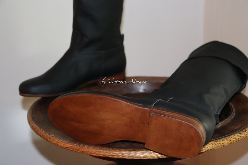 May include: A pair of black leather boots with brown wooden soles. The boots are on a wooden surface with a woven pattern.