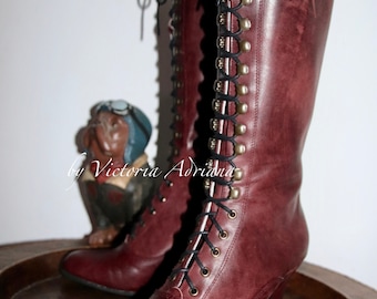 May include: Burgundy leather boots with black laces and brass eyelets. The boots have a chunky heel and a lace-up front. The boots are sitting on a wooden surface.