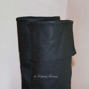 May include: A pair of black over-the-knee boots with a fold-over top. The boots are made of a smooth, faux leather material. The text "by Victoria Adriana" is visible on the boots.