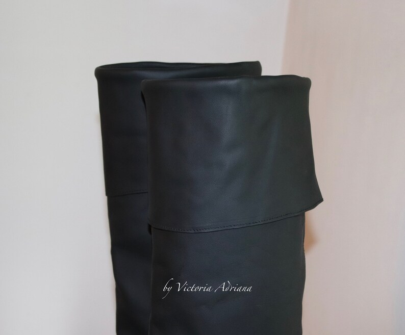May include: A pair of black over-the-knee boots with a fold-over top. The boots are made of a smooth, faux leather material. The text "by Victoria Adriana" is visible on the boots.