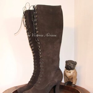 May include: Brown suede knee-high boots with lace-up detail and a block heel. The boots are on a wooden surface with a small decorative dog figurine.