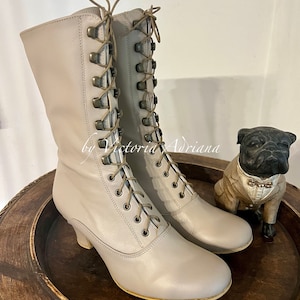 Pode incluir: Um par de botas de couro cinza claro com cadar&ccedil;os e salto baixo. As botas t&ecirc;m um detalhe de cadar&ccedil;os decorativos no eixo e est&atilde;o sobre uma superf&iacute;cie de madeira.