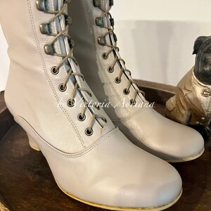 Pode incluir: Um par de botas de couro cinza claro com cadar&ccedil;os na frente e um salto pequeno. As botas t&ecirc;m um detalhe de costura decorativa em torno do bico e do calcanhar.