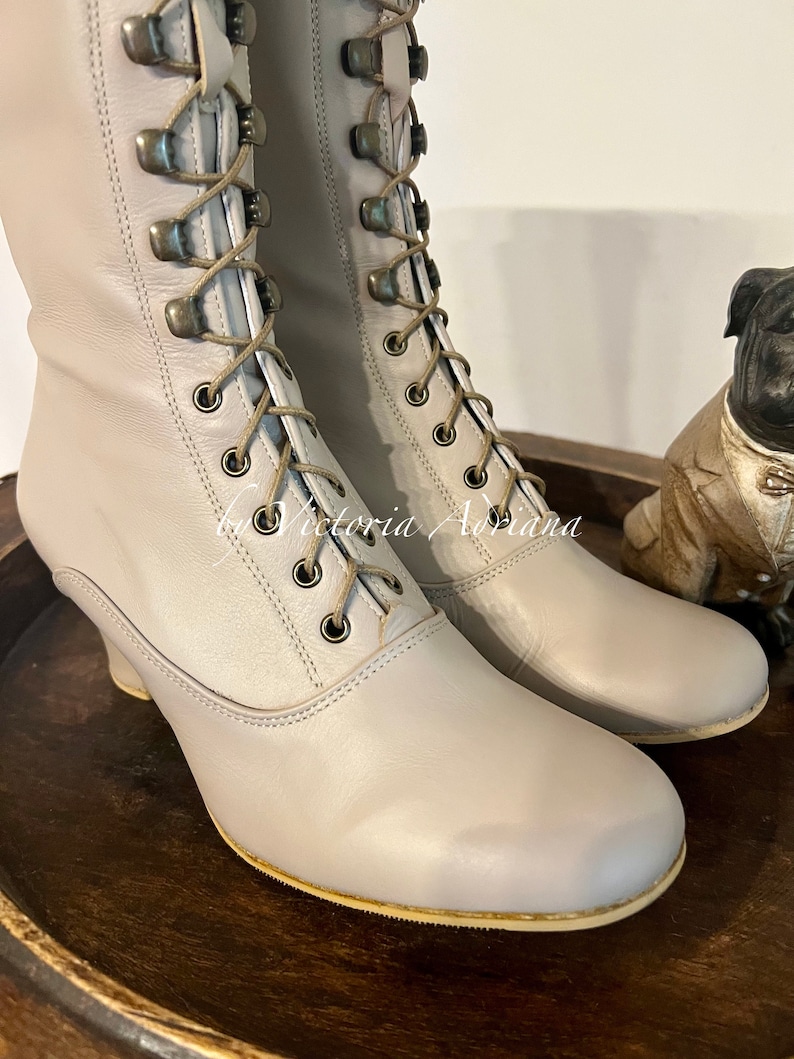 Pode incluir: Um par de botas de couro cinza claro com cadar&ccedil;os na frente e um salto pequeno. As botas t&ecirc;m um detalhe de costura decorativa em torno do bico e do calcanhar.