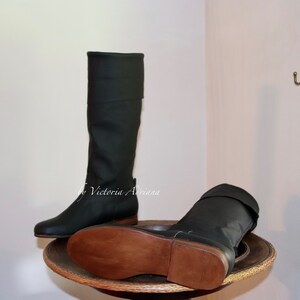 May include: A pair of dark green leather boots with brown leather soles. The boots are tall and have a fold-over top. The boots are on a wooden stand.