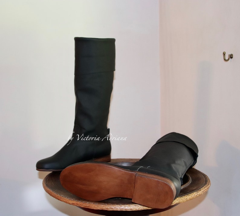 May include: A pair of dark green leather boots with brown leather soles. The boots are tall and have a fold-over top. The boots are on a wooden stand.