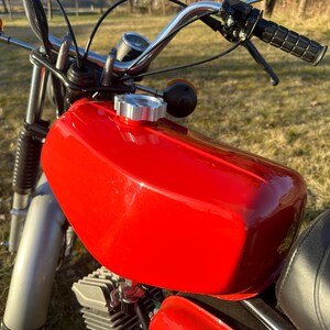 Op de afbeelding: Een rode motorfiets benzinetank met een zilveren benzine dop. De tank is glanzend en heeft een lichte kromming.