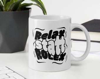 May include: White ceramic mug with the words "Relax, Start, Rolex" in a black, bubble-style font. The mug is on a white surface with a plant and books in the background. A keyboard is in the foreground.