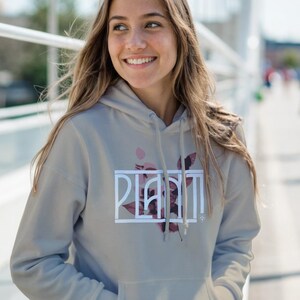 Könnte beinhalten: Ein hellgrauer Kapuzenpullover mit dem Wort "PLANT" in einem weißen rechteckigen Rahmen und einem floralen Design. Der Kapuzenpullover hat eine Vordertasche und eine Kapuze mit Kordelzug. Die Person trägt eine blaue Jeans.