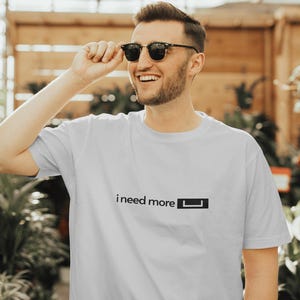 May include: A man wearing sunglasses is smiling and looking to the right. He is wearing a light grey t-shirt with the text "i need more └" printed on the front.