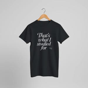 Op de afbeelding: Zwart T-shirt met de tekst "That's what I studied for" in witte letters. Het shirt hangt aan een houten hanger tegen een lichtgrijze achtergrond. De tekst is in een stijlvol lettertype.
