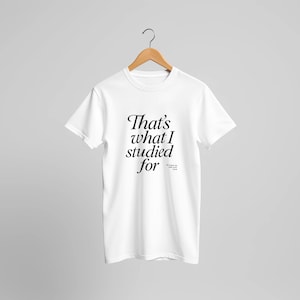 Op de afbeelding: Wit T-shirt met de tekst "That's what I studied for" in zwart script. Het shirt hangt aan een houten hanger tegen een lichtgrijze achtergrond. De tekst is in een stijlvol lettertype.
