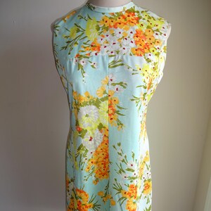 May include: A vintage sleeveless dress with a high neckline. The dress features a light blue background with a vibrant floral pattern in yellow, orange, and white. The dress is knee-length and appears to be made of a lightweight fabric.