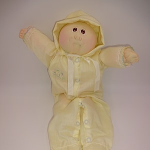 Può includere: Una bambola Cabbage Patch Kid gialla vestita con una tutina gialla con cappuccio, bottoni bianchi e un nastro bianco. La bambola ha un viso dipinto con occhi marroni e un incarnato rosato.