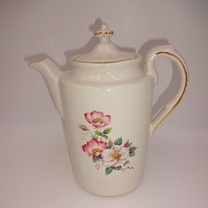 Op de afbeelding: Crèmekleurige keramische theepot met een bloemenontwerp van roze en witte bloemen met gele centra. De theepot heeft een deksel, een gebogen handvat met gouden rand en een tuit. Vintage stijl.