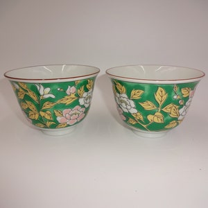 Può includere: Due tazze da tè in ceramica bianca con motivi floreali verdi. Le tazze hanno un bordo rosso e un interno bianco.
