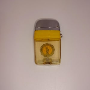 Może przedstawiać: Zapalniczka w stylu vintage ze srebrną górą i żółtym paskiem. Przezroczysty korpus posiada złoty emblemat z Statuą Wolności. Klasyczny przedmiot kolekcjonerski.