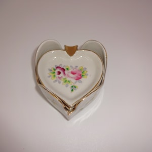 以下が含まれることがあります： 金色の縁取りが施された白いハート型のセラミック製灰皿が3つセットになっています。中央の灰皿には、ピンクと黄色のバラの花柄が描かれています。