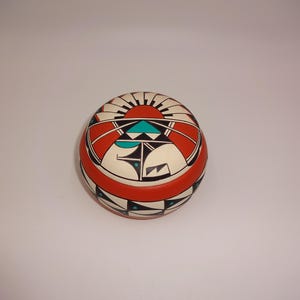 May include: A round ceramic lidded jar featuring a matte finish with a geometric pattern in red, black, white, and turquoise. The lid displays a sunburst motif, while the base is decorated with repeating triangular and linear black and white shapes.