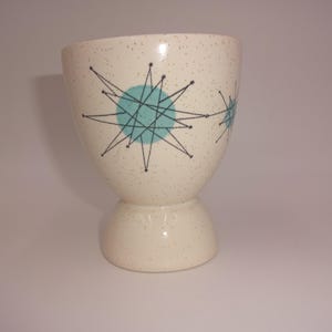 May include: A cream-colored ceramic cup with a speckled finish and a pedestal base. The exterior features a graphic pattern of black starburst lines layered over solid light blue circles, creating a repeating geometric design around the vessel.