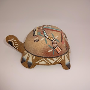 May include: A ceramic turtle figurine with a textured tan surface. The shell features painted geometric patterns in red, black, white, and blue. The head and legs are decorated with white and black feather-like motifs on a speckled brown base.