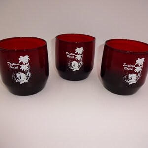 May include: Three red-tinted glass tumblers with a dark base. Each tumbler features a white graphic of palm trees, a wave, and the words "Daytona Beach". The glasses are cylindrical with a slightly angled design.