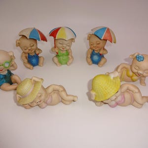 May include: A collection of ceramic pig figurines in beach attire. Some pigs wear sunglasses, swimsuits, and hats. Others hold colorful umbrellas. The figurines are in various poses, suggesting a relaxing day at the beach.