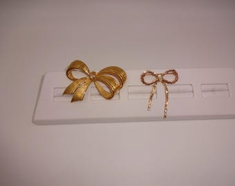 May include: Two gold-toned bow brooches. The larger brooch has a textured surface and a small, clear gem at the center. The smaller brooch has a twisted rope design with a gem at the center. Both are displayed on a white surface.