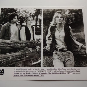 May include: A black and white photograph of a couple standing by a wooden fence. The woman is wearing a waistcoat and the man is wearing a long-sleeved shirt. The photo is from the USA Network television show "Wounded Heart".