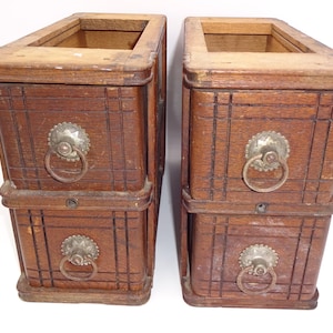 May include: Two antique wooden storage boxes with a dark brown finish. Each box has two drawers with decorative metal ring pulls and a square opening at the top. The boxes have a grid-like pattern carved into the wood.