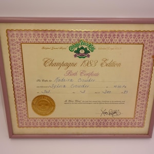 Puede incluir: Certificado de nacimiento enmarcado de Cabbage Patch Kids "Champagne 1983 Edition". El certificado está en un marco rosa con un borde dorado y presenta el nombre "Madeira Crowder". El certificado está fechado el 3 de diciembre de 1983.