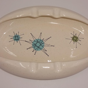 May include: An oval-shaped ceramic ashtray with a cream-colored speckled glaze. The interior features three black starburst patterns with blue and green circular accents. The rim includes two notches for resting items, creating a functional design.