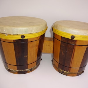 May include: A pair of wooden bongo drums. The drums have light tan drumheads, yellow trim, and are constructed with alternating vertical stripes of light and dark brown wood. The drums are connected by a small wooden block.