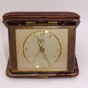 May include: A vintage travel alarm clock in a brown leather case. The clock face is cream-colored with gold-tone hands and numerals. The case is rectangular and folds open. The clock measures approximately 3 inches wide.