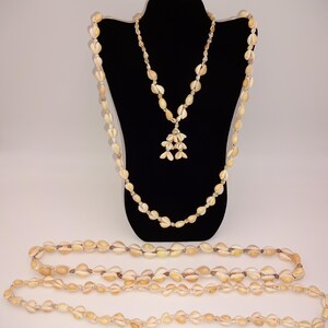 Peut inclure: Trois colliers faits de coquillages beiges. Un collier est présenté sur un mannequin noir, avec un pendentif de coquillages groupés. Les deux autres colliers sont disposés, mettant en valeur la couleur et la texture naturelles des coquillages.