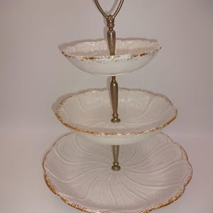 May include: A three-tiered serving stand with a white ceramic base and gold trim. Each tier has a scalloped edge and a delicate embossed design. The stand has a metal handle for easy carrying.