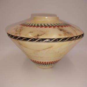May include: A cream-coloured wooden bowl with a wide, rounded shape. It features a black wavy band and bands of red and teal geometric patterns. The wood grain is visible, and the bowl has a smooth, polished finish. A decorative item.
