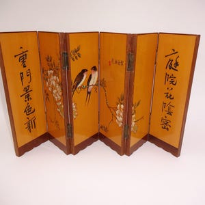 Puede incluir: Un biombo plegable de madera con un acabado ámbar cálido. Los paneles presentan pinturas de pájaros, flores y caligrafía china. El biombo está articulado y se mantiene en posición vertical, mostrando su diseño decorativo.
