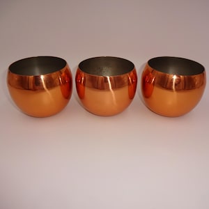May include: Three copper shot glasses, each with a rounded, slightly flared rim. The glasses are shiny and reflect light.