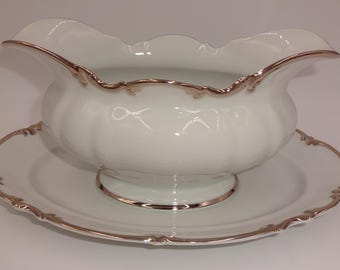 May include: A white porcelain gravy boat with a scalloped rim and a matching oval underplate. Both pieces feature a metallic silver trim along the edges. The gravy boat has a curved handle and a wide pouring spout, resting on a pedestal base.