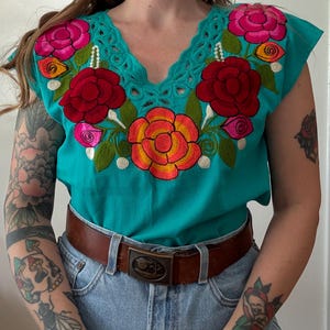 以下が含まれることがあります： Vネックとキャップスリーブのターコイズ刺繍トップス。赤、ピンク、オレンジ、黄色の鮮やかな花の刺繍が施されています。装飾的なバックルが付いた茶色の革のベルトは、青いデニムジーンズと一緒に着用されています。