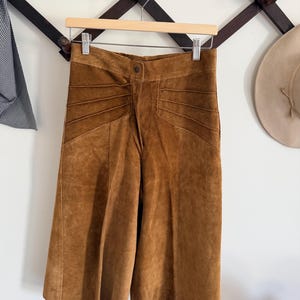 May include: High-waisted brown suede pants featuring a wide-leg silhouette and a structured waistband. The front of the garment displays decorative horizontal stitched pleats extending from the side seams toward the center, with a single button closure.