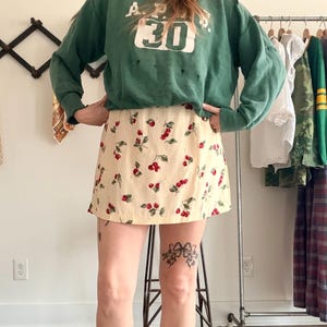 May include: A cream-colored mini skirt featuring a repeating pattern of red cherries with green leaves, paired with a long-sleeved forest green sweatshirt that displays the white text A.P.S. and the number 30 on the front chest area.