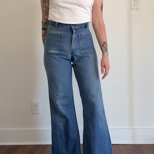 Könnte beinhalten: Hoch taillierte blaue Jeans aus Denim mit weitem Bein und zwei großen, quadratischen aufgesetzten Taschen auf der Vorderseite. Die Jeans verfügt über einen klassischen Knopf- und Reißverschluss, Gürtelschlaufen und eine mittelblaue Waschung.