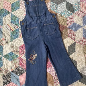 May include: Blue denim overalls with a cowboy hat embroidered on the left leg. The overalls have adjustable straps and two front pockets.