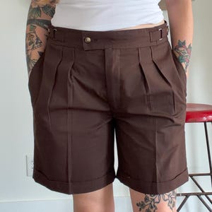 May include: Dark brown tailored shorts featuring a high-waisted design with double pleats, a button closure, and adjustable side tabs with metal buckles. The shorts have a structured waistband, side pockets, and cuffed hems that fall above the knee.