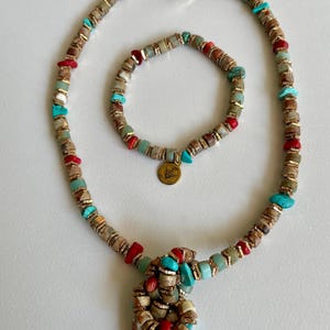 Könnte beinhalten: Ein Halsketten- und Armband-Set mit einer Mischung aus farbigen Perlen. Die Halskette und das Armband bestehen aus braunen, roten, türkisfarbenen und goldfarbenen Perlen. Die Halskette hat einen einzigartigen Anhänger aus geclusterten Perlen.
