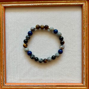 Könnte beinhalten: Ein Armband mit Perlen aus verschiedenen Steinen in Blau-, Grau-, Braun- und Schwarztönen. Das Armband liegt auf einer weißen Oberfläche in einem orangefarbenen Holzrahmen. Die Perlen haben einen Durchmesser von etwa 0,8 cm.