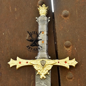 May include: A decorative sword with a gold-colored crown and crossguard, featuring red accents. The handle is silver with gold details and a logo. The blade has a patterned design. The sword is displayed against a brown background.