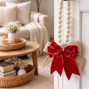 May include: A decorative door hanger featuring a red satin bow and burlap accents, suspended by a strand of wooden beads. The door hanger is attached to a white door with a black handle. The background shows a living room with a sofa and coffee table.